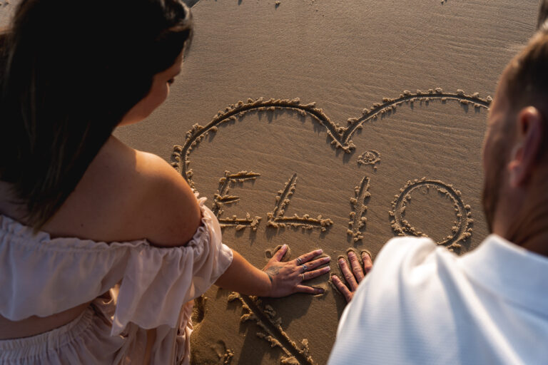 Coeur avec prénom d'un bébé noté dans le sable avec les mains des parents dessous lors de leur séance photo grossesse sur une plage du bassin d'arcachon