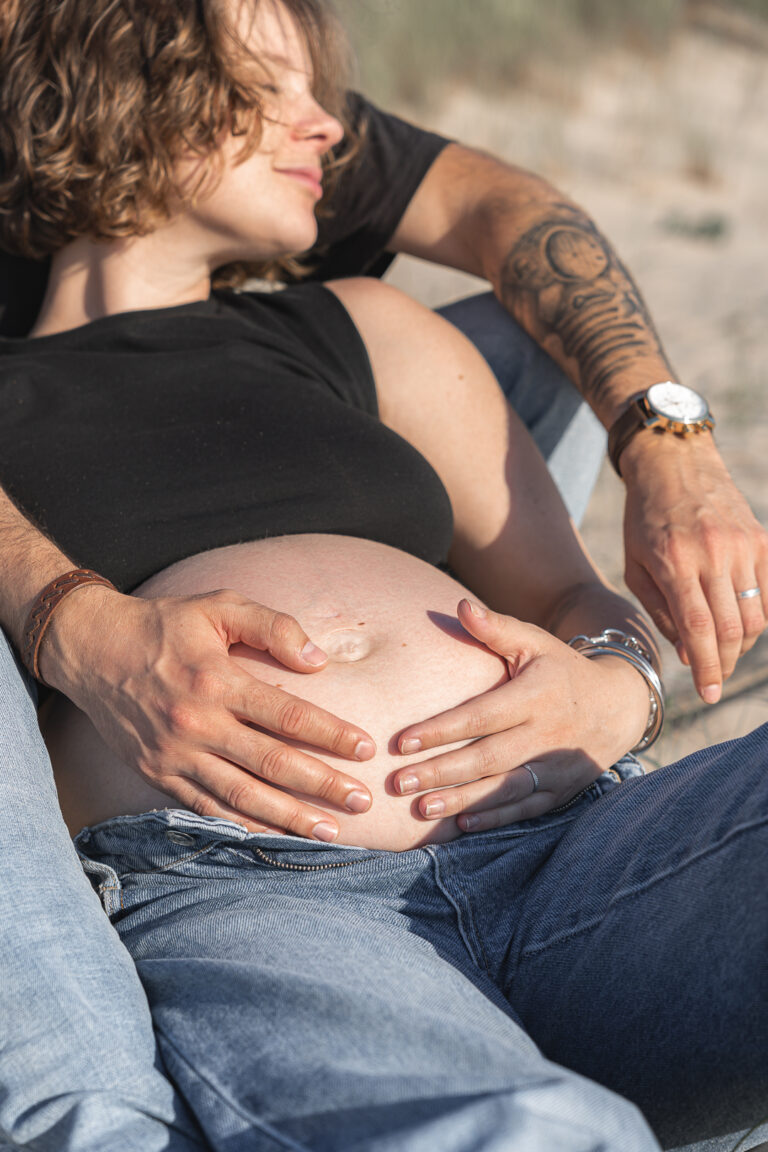 Ventre d'une femme enceinte allongée entouré par les mains du papa et de la maman lors de leur séance photo grossesse aux dunes du pilat sur le bassin d'arcachon