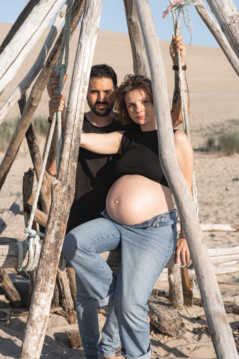 futurs parents qui regardent l'objectif assis dans une structure en bois des dunes du pilat lors de leur séance photo grossesse sur le bassin d'arcachon