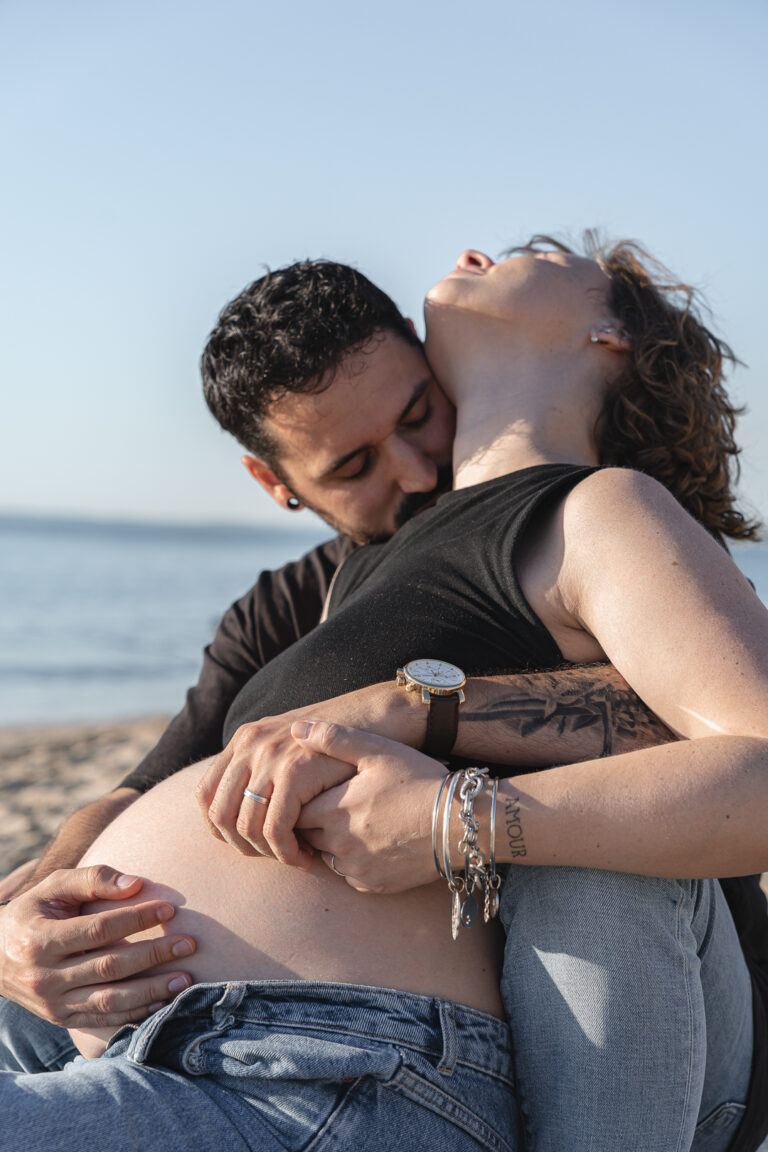Futur papa qui embrasse la future maman dans le cou avec sensualité pendant leur séance photo grossesse à la plage de la dune du pilat