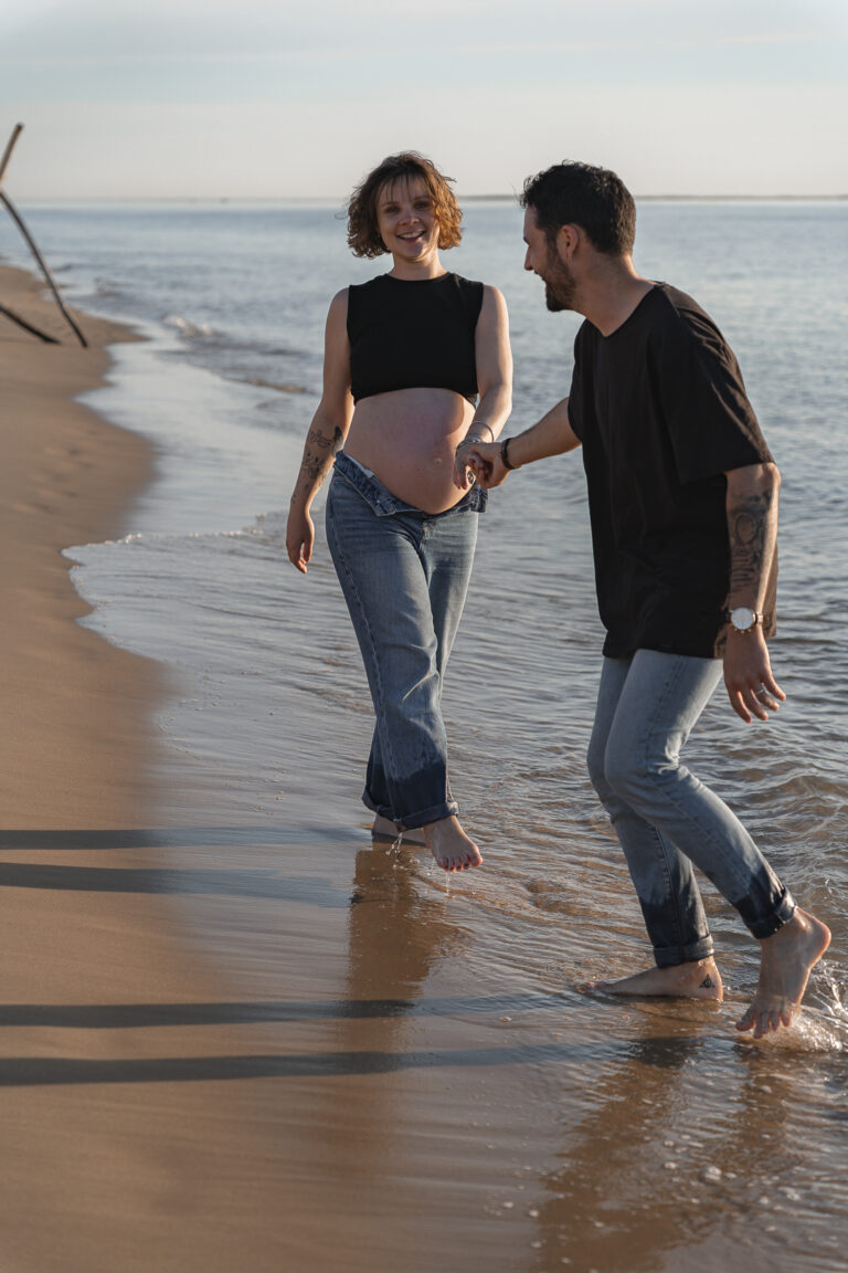 Futurs parents qui marchent sur le sable, les pieds dans l'eau en riant lors de leur séance photo grossesse sur le bassin d'arcachon