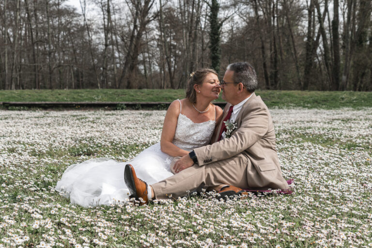Mariés qui se regardent dans les yeux assis par terre dans un champs de fleur près d'un bois dans la Beauce par photographe mariage eure-et-loir durant la séance couple d'un mariage civil