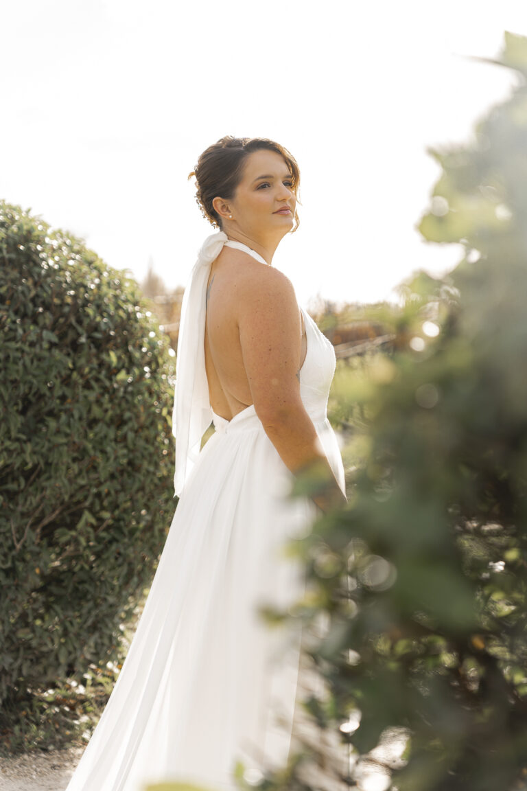 Mariée qui regarde au loin, cachée par des buissons des vignobles lors du portrait de la mariée au coucher du soleil de la cérémonie laïque de son mariage au chateau de courlat à Lussac